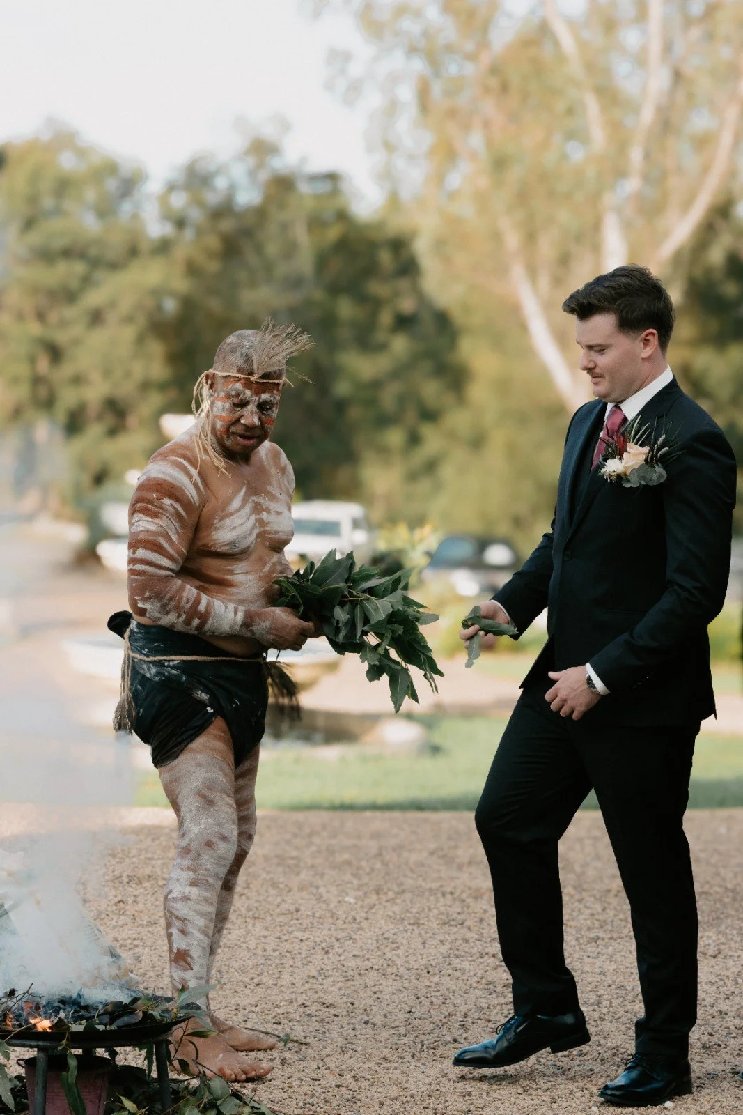 A Indigenous man with traditional paint, wearing a loincloth, handing a plant to a man in a black suit at an outdoor ceremony.