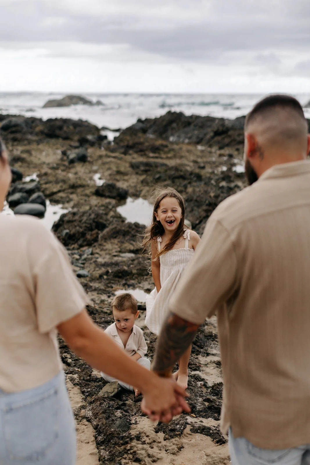 A family of four holding hands on a rocky beach with the ocean and cloudy sky in the background. The children are smiling and laughing.