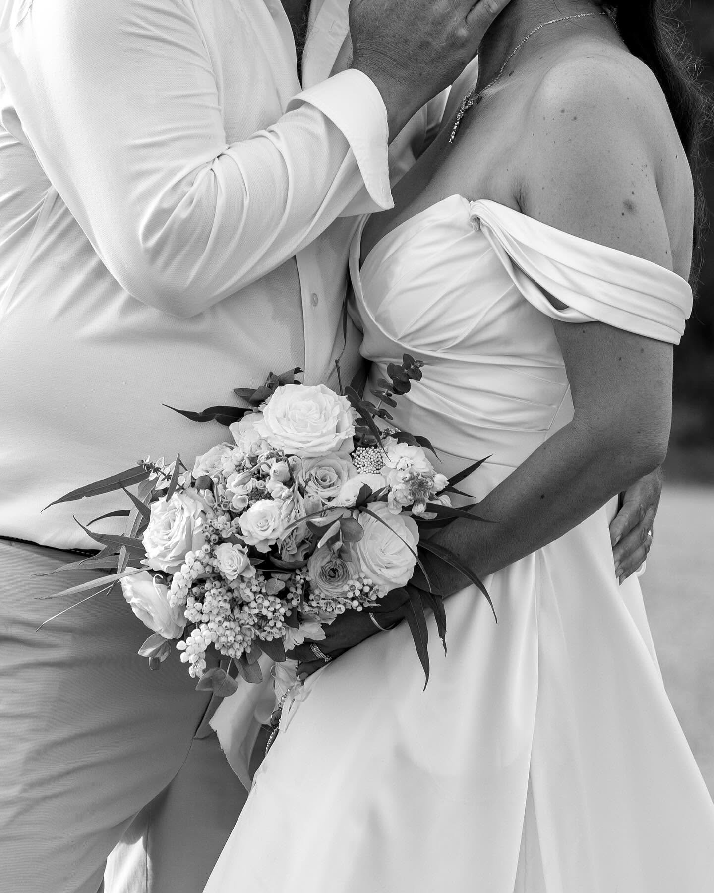 A bride in an off-shoulder wedding dress holding a large bouquet of flowers, kissing a man dressed in a white shirt, at a wedding.