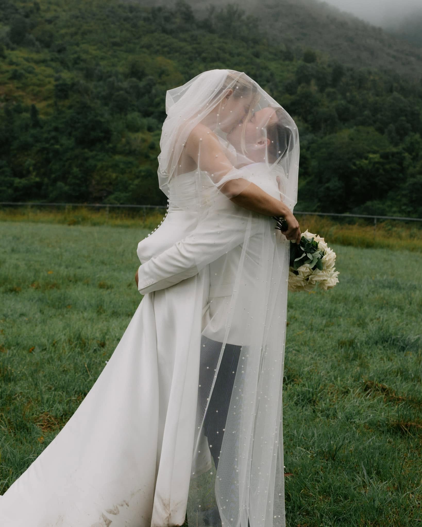 A bride in a wedding gown with a long veil holding a bouquet of white roses outdoors in a grassy field with green hills and misty mountains in the background.