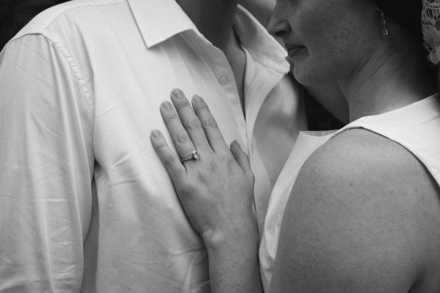 A close-up of a couple in an intimate moment, with the woman's hand resting on the man's chest, showing an engagement ring.