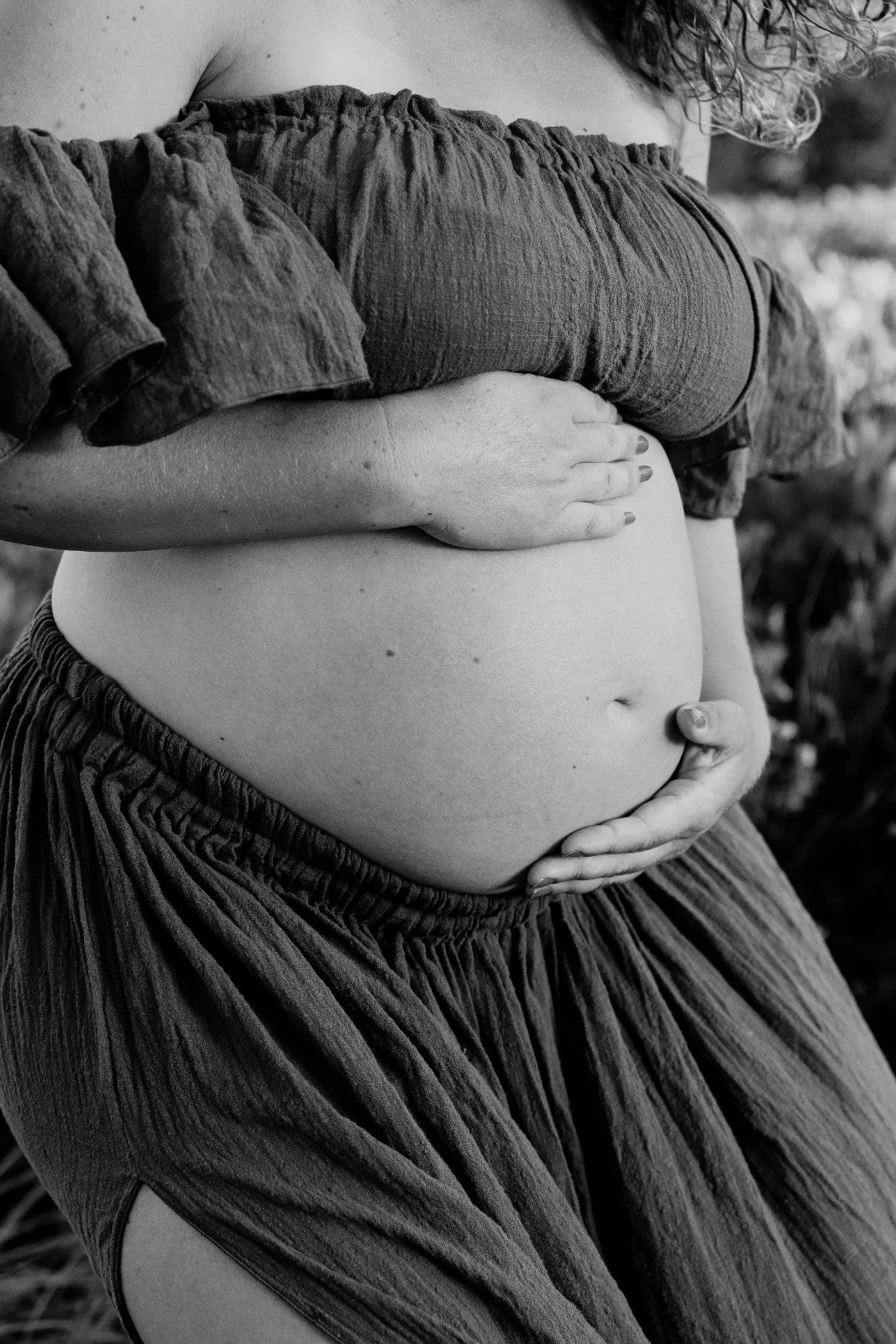 Close-up black and white photo of a pregnant woman’s belly, touching it with both hands, wearing a loose dress with ruffled sleeves.