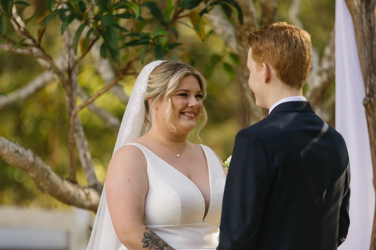 A bride and groom exchanging vows outdoors, smiling at each other during their wedding ceremony surrounded by trees.