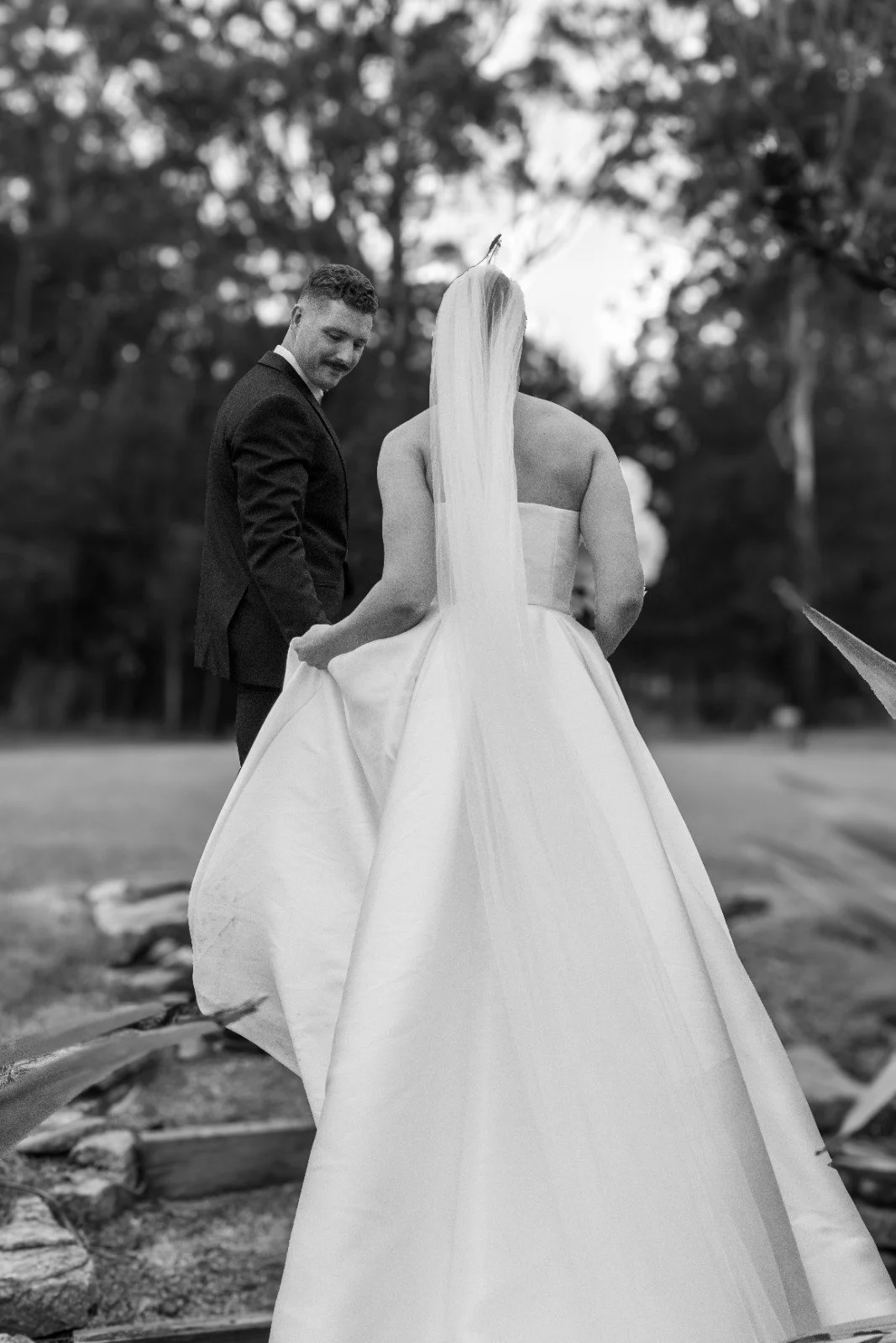 A black and white photo of a bride in a strapless wedding gown and veil holding her dress, standing outside on a pathway with trees in the background, while a groom in a tuxedo looks at her.