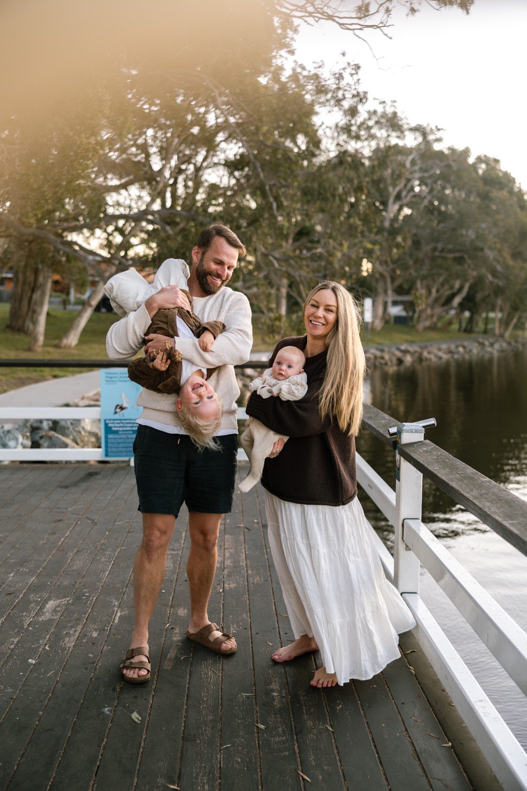 A happy family of four standing on a wooden pier by a river, with trees in the background during daytime. The father is holding a young boy upside down, and the mother is holding a baby girl, all smiling and enjoying their time together.