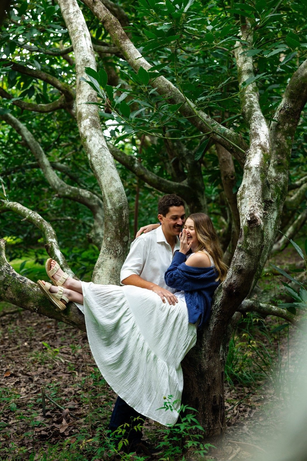 A couple sitting on a tree branch in a green forest, laughing and enjoying each other's company.