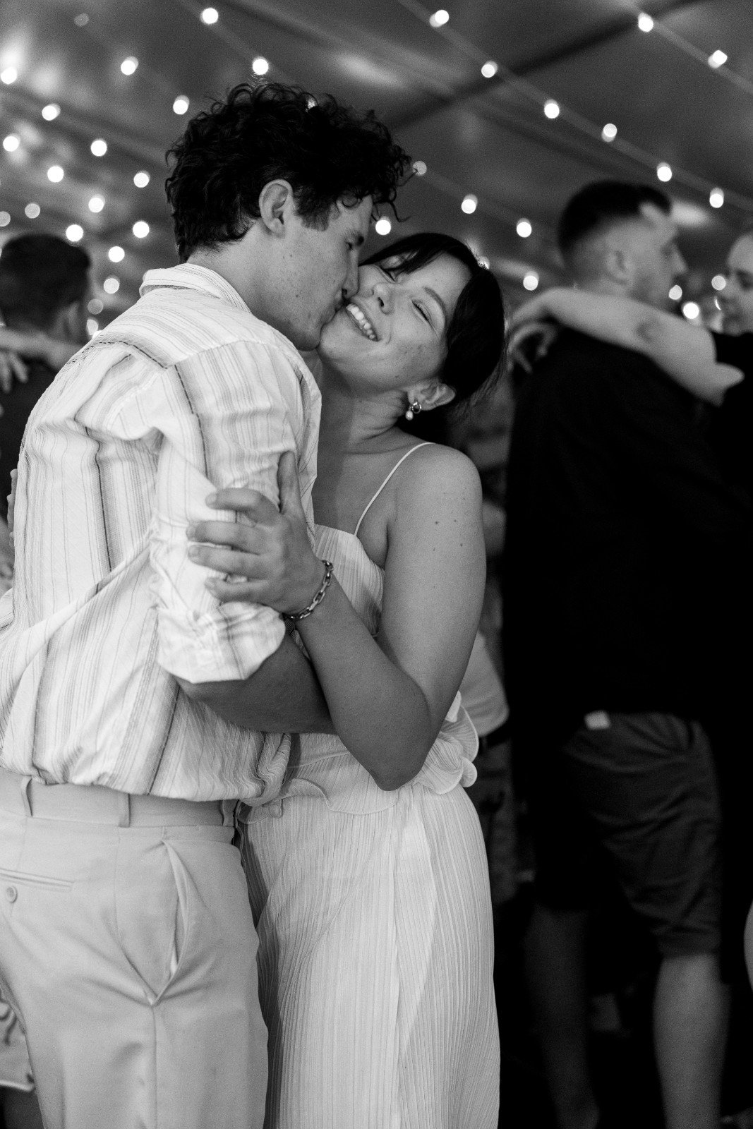 A black and white photo of a young couple dancing closely at a celebration, with string lights overhead and other couples in the background.