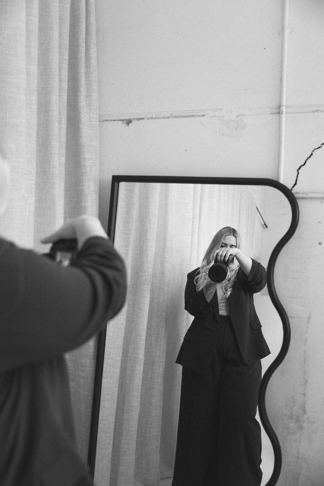A woman with long blonde hair taking a photo of herself in a mirror. She is wearing a blazer and smiling. The mirror has an irregular, curved shape. The background shows a wall with white paint and some cracks, along with a curtain on the left.