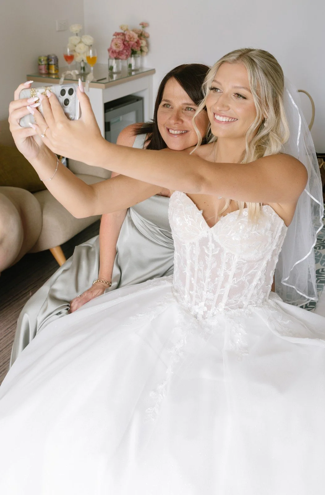 A bride and a woman, possibly her mother, taking a selfie together in a hotel room, with flowers and drinks in the background.