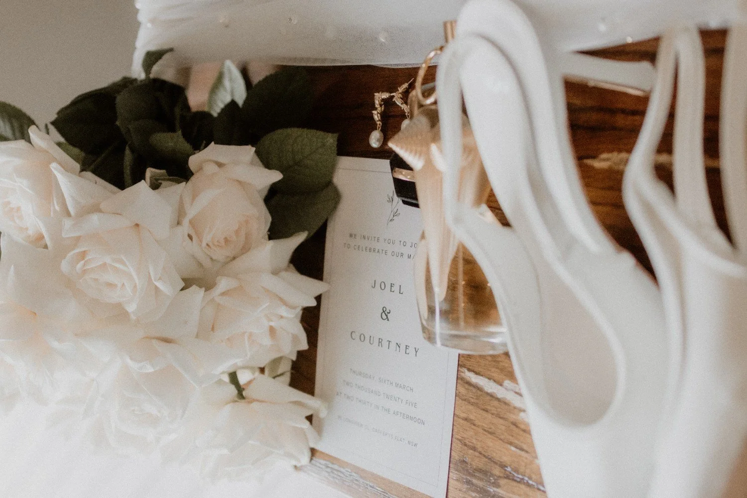 Close-up of wedding decor with white flowers, a wine glass, jewelry, and an invitation for Joel and Courtney on a wooden surface.