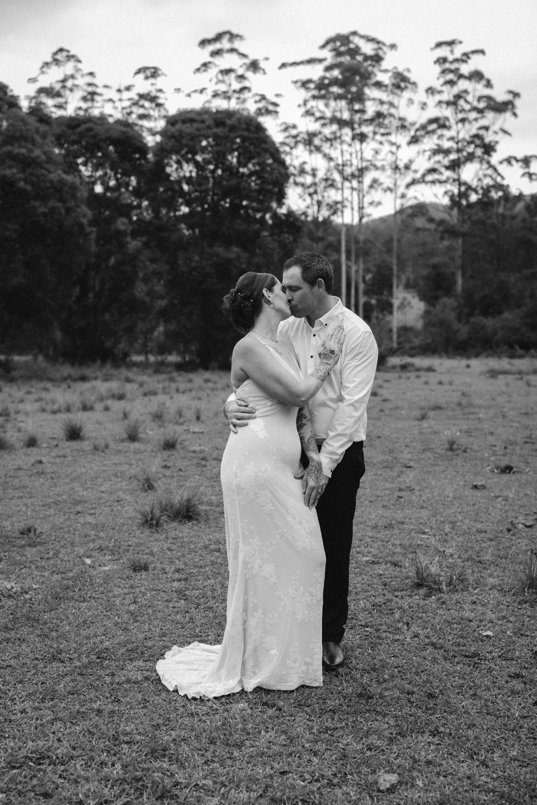 A black and white photo of a couple kissing outdoors. The woman is wearing a white wedding dress with floral details, and the man is in a white shirt and dark pants. They are standing on grass with trees and hills in the background.