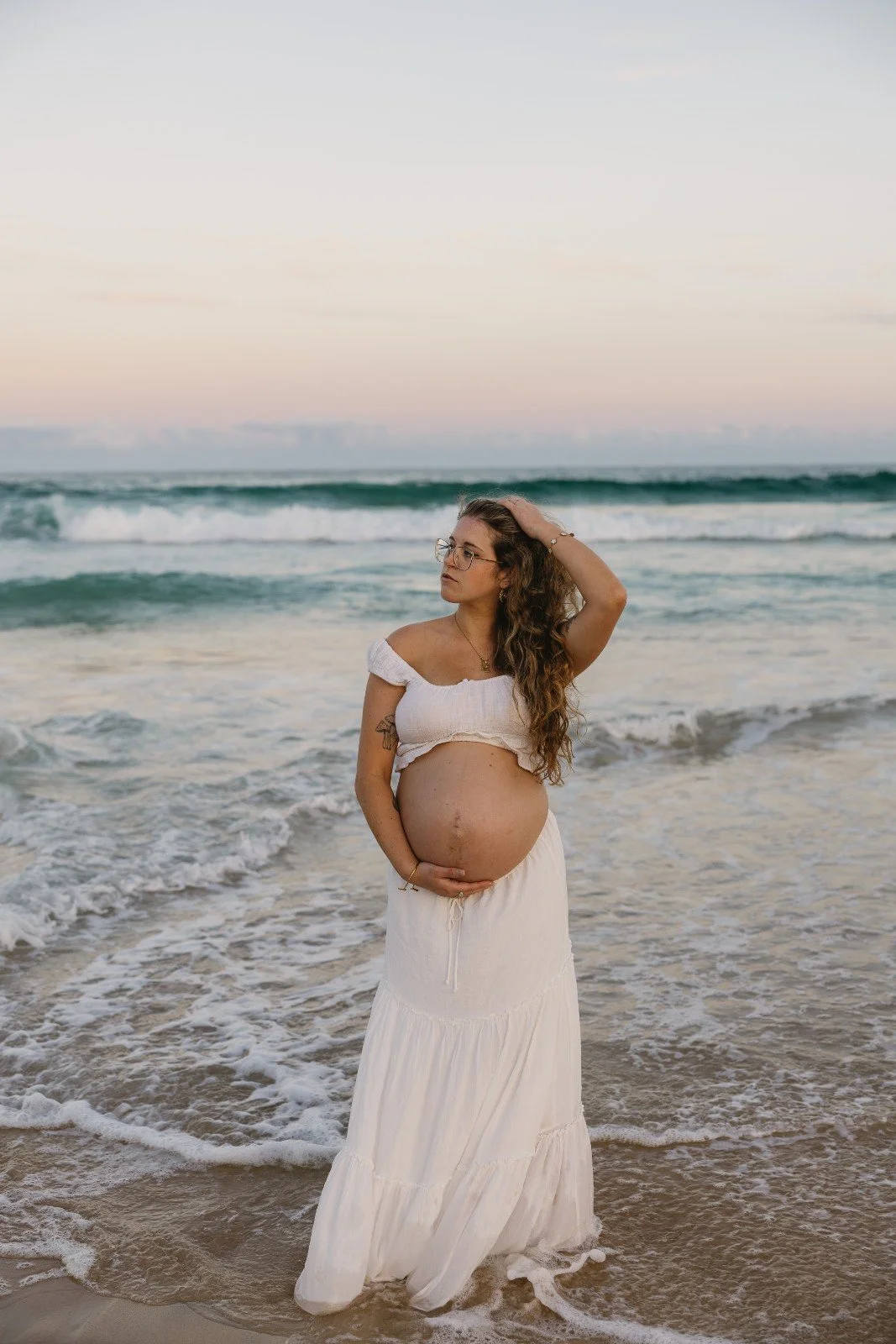 Pregnant woman standing in the ocean at sunset, wearing a white off-shoulder top and a long skirt, with one hand on her belly and the other touching her hair.