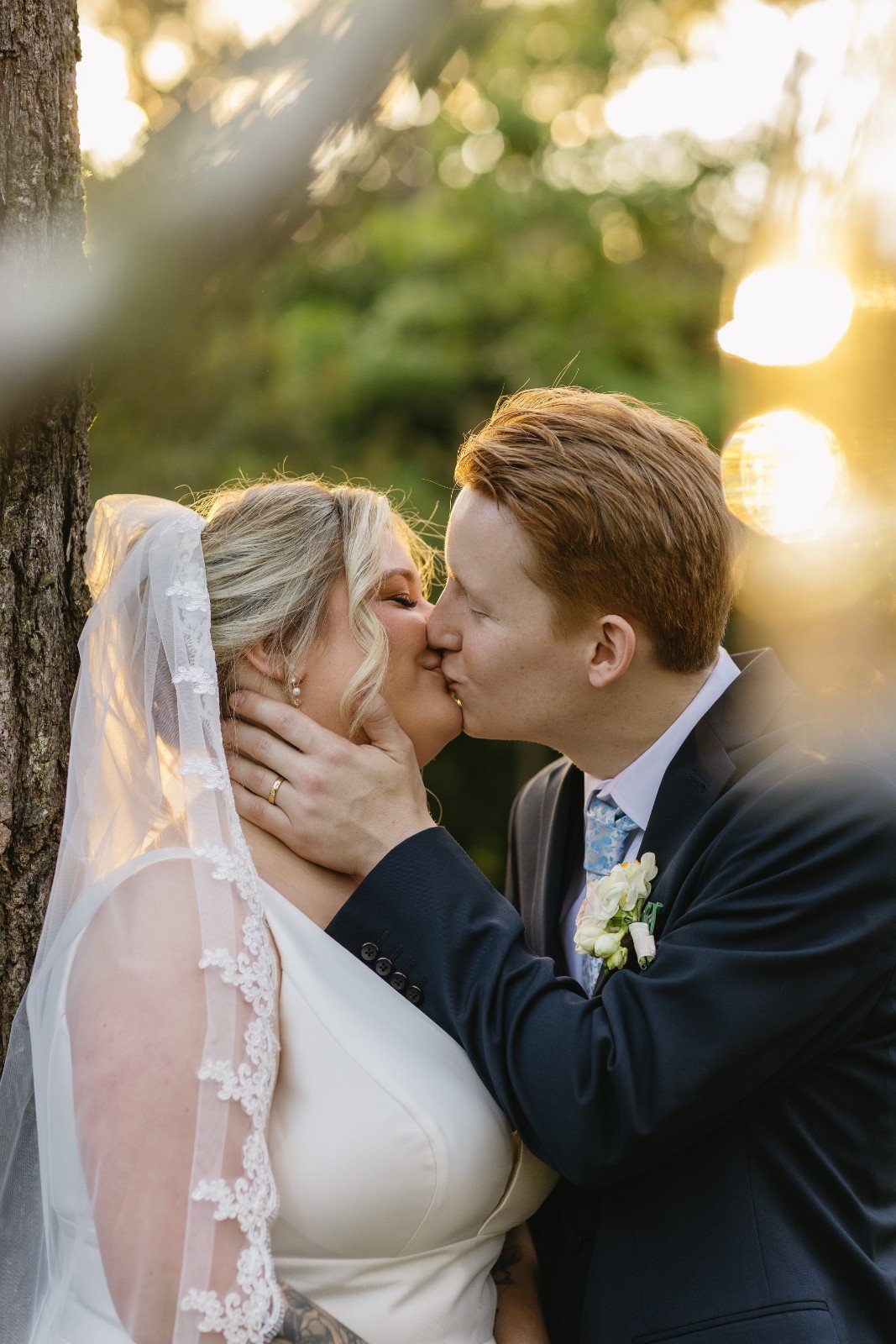 A bride and groom kiss outdoors during sunset, with blurred greenery and sunlight in the background.