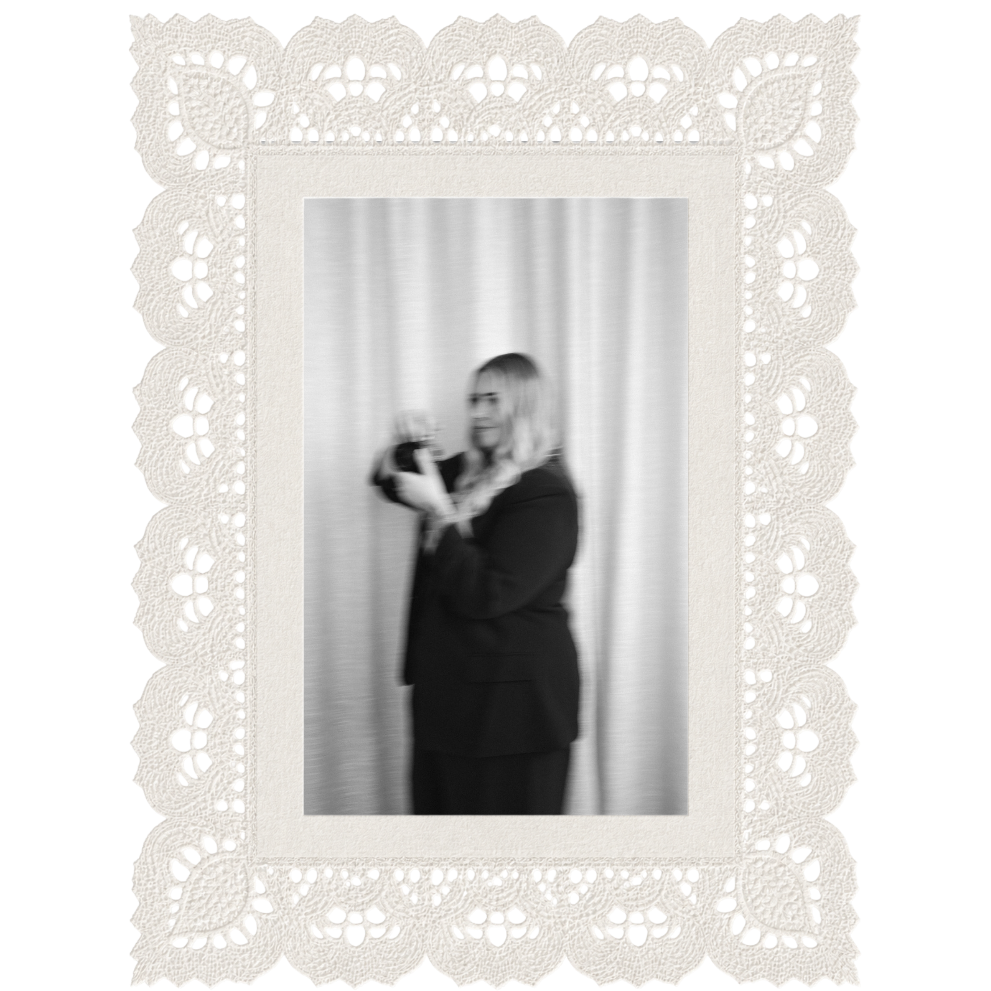 The image shows a black and white photograph of a woman with long blonde hair, wearing glasses and a dark top, standing in front of a light-colored, textured wall or curtain.