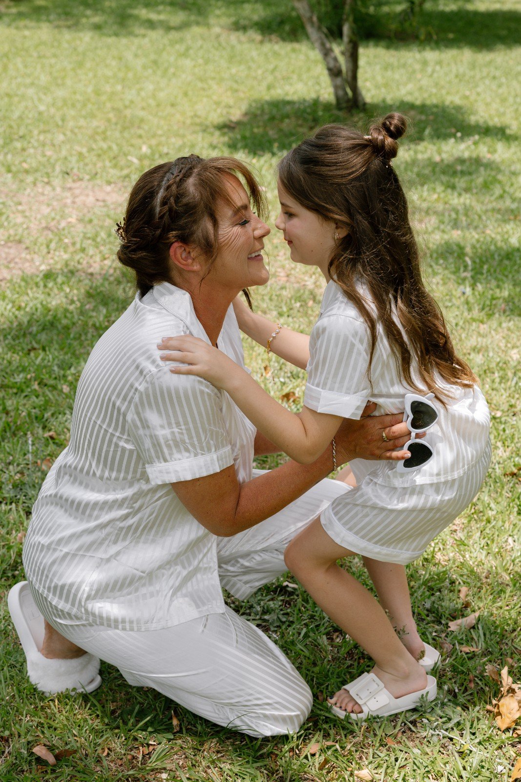 A woman and young girl are smiling and hugging outdoors on grass, both wearing white striped pajamas.