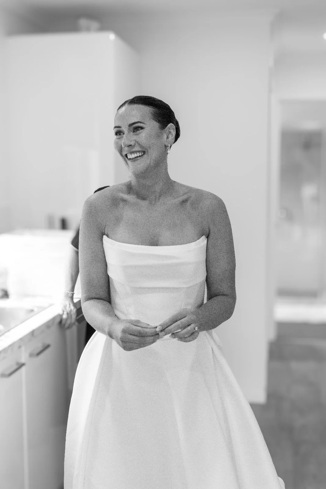 A woman in a strapless wedding gown smiling in a modern kitchen, with her hair pulled back and wearing earrings.