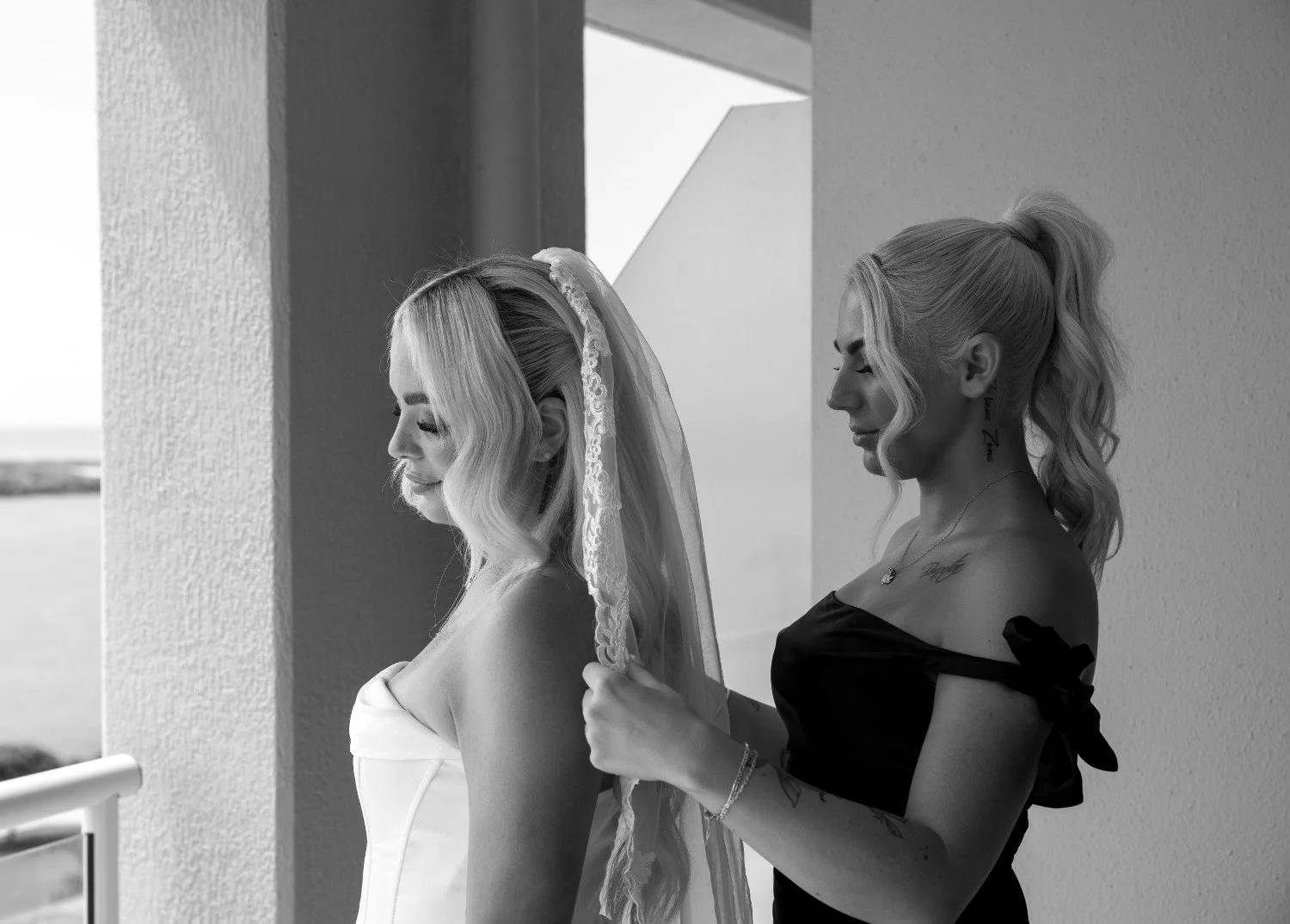 A bride with long wavy blonde hair and a veil standing on a balcony, as a woman in a black dress adjusts the veil.