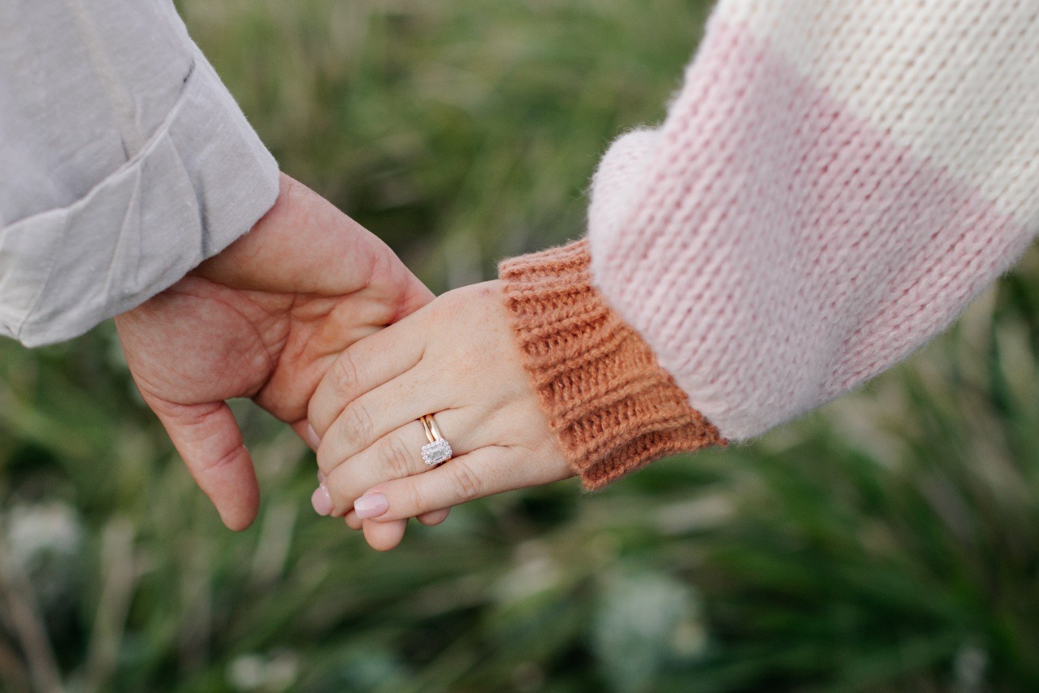 A close-up of two people holding hands, showing an engagement ring on the woman's finger. The woman wears a pink and orange sweater, and the man wears a light-colored shirt. The background appears to be outdoors with greenery.