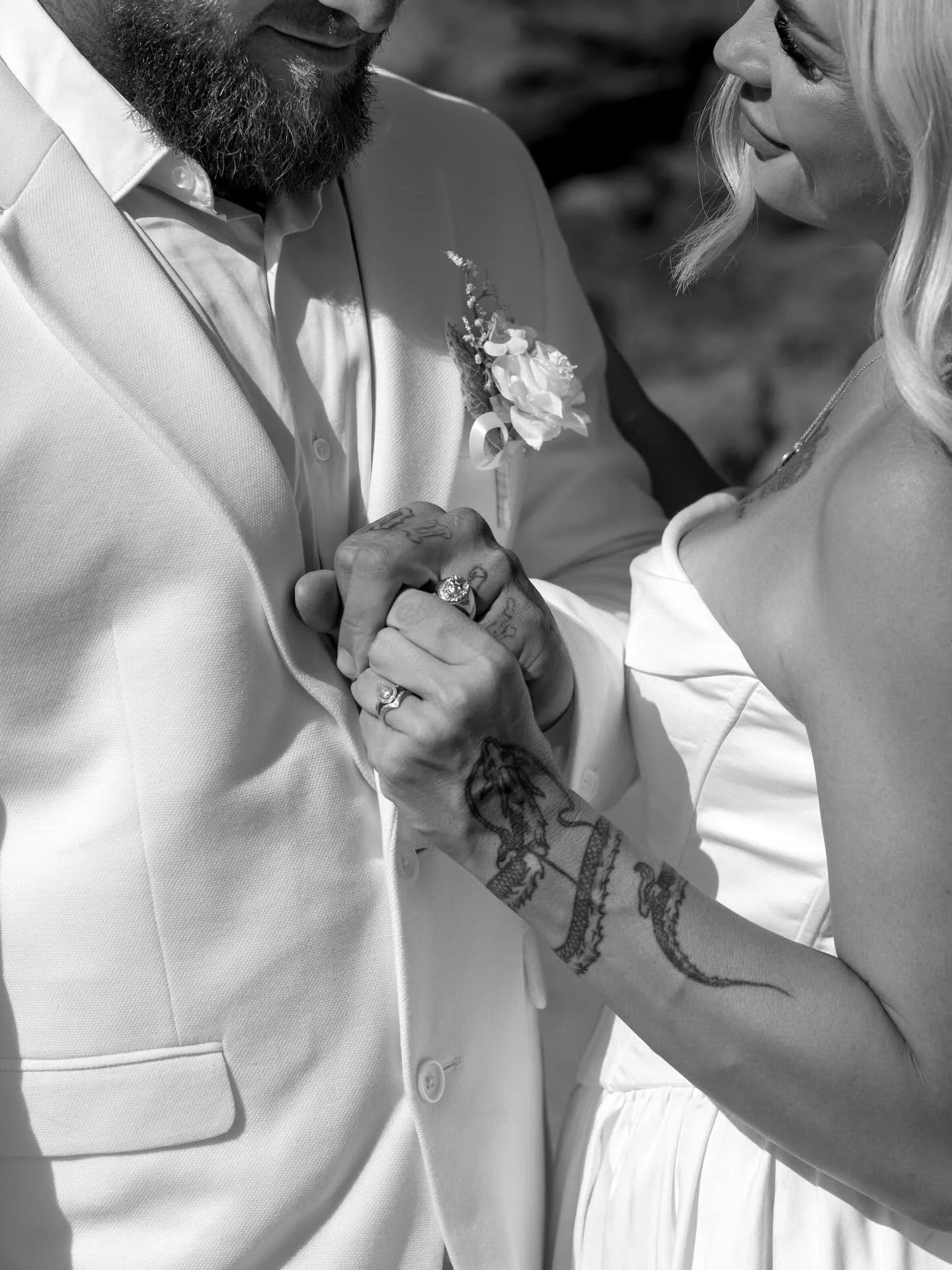 A close-up of a couple holding hands during their wedding, focusing on their intertwined fingers and wedding rings, with the woman's tattooed arm visible.