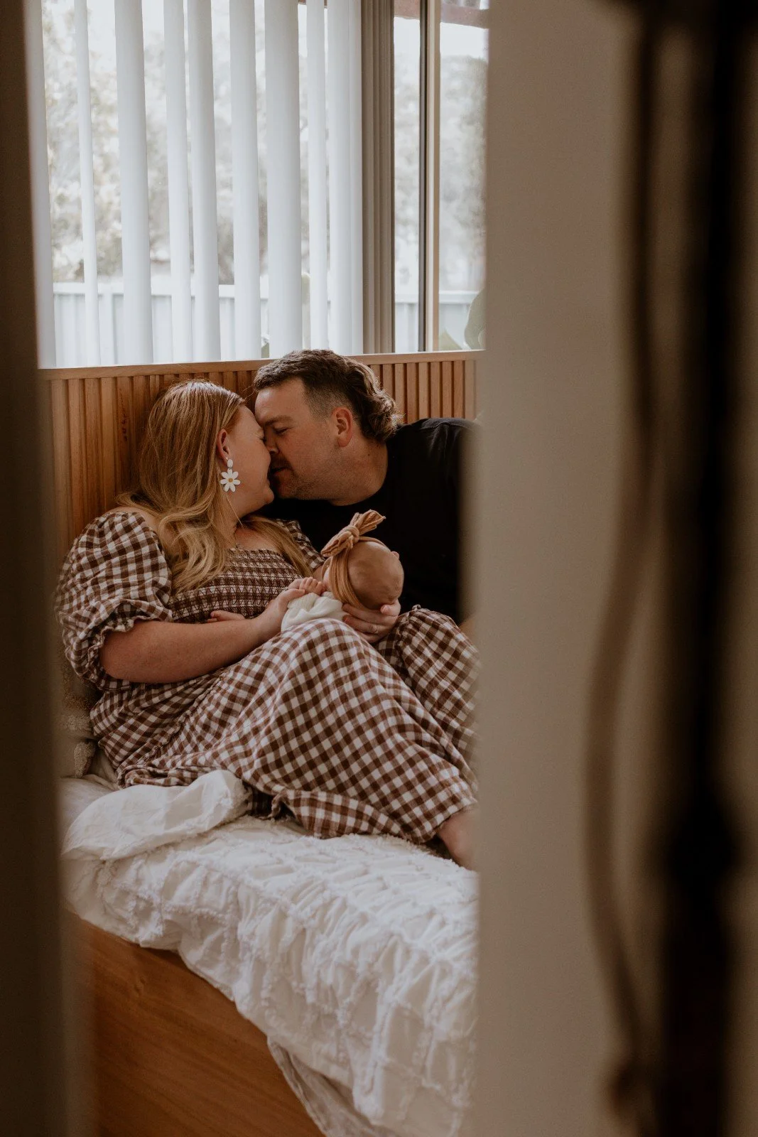 A family of three, a woman, a man, and a baby, sitting on a bed in a cozy room, sharing a tender moment with their foreheads touching, the woman holding the baby in her arms.