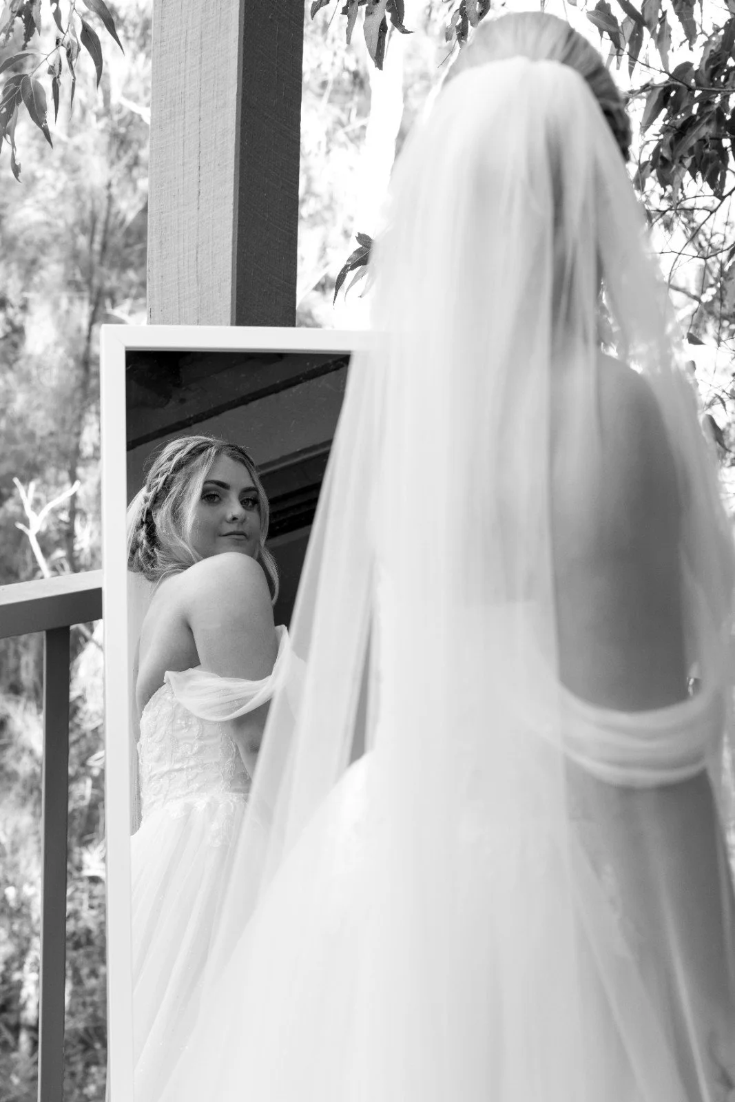 A woman in a wedding dress looking at her reflection in a mirror outdoors with trees and foliage surrounding her.