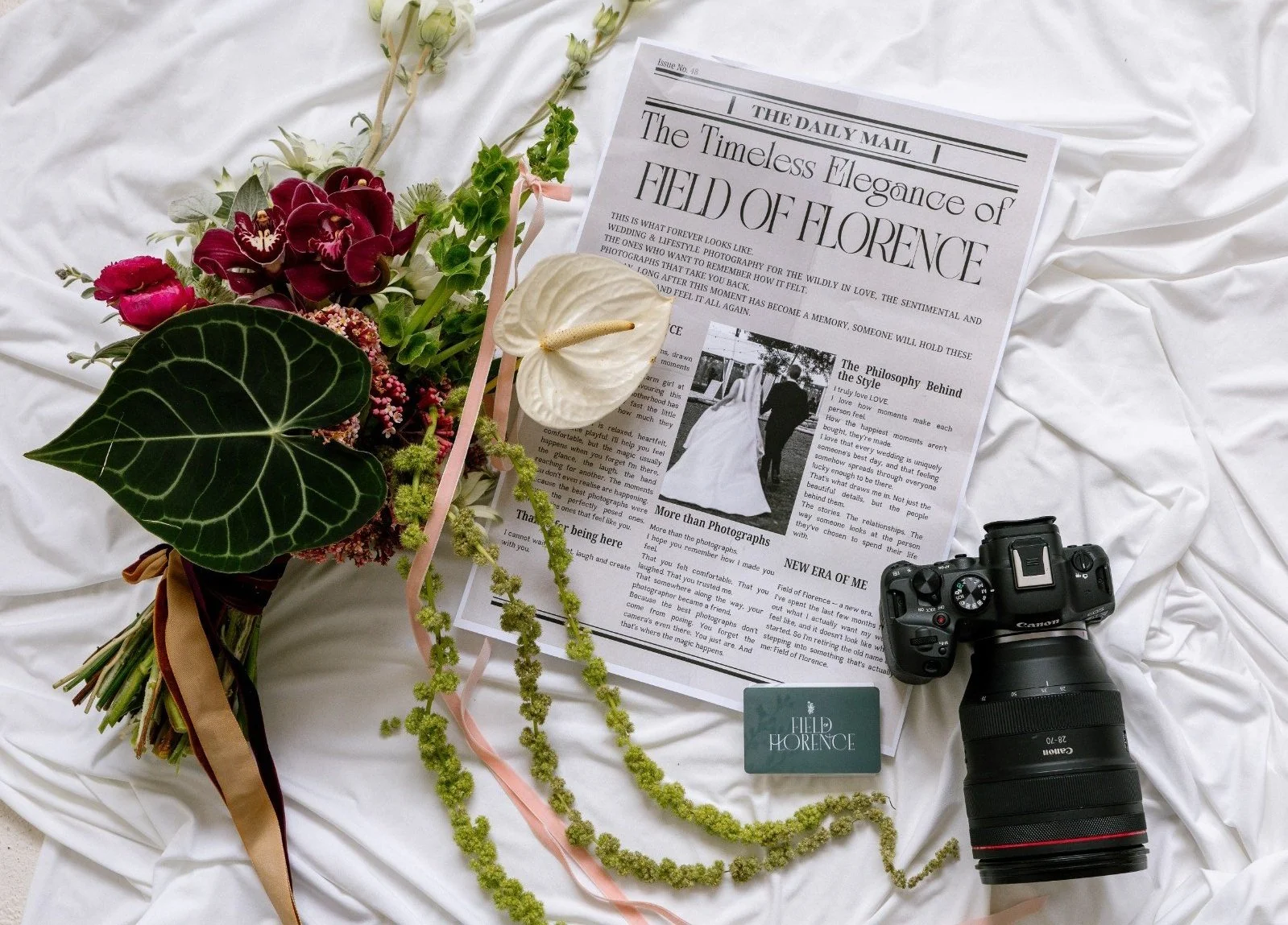 A bouquet of flowers, a photo of a wedding couple, a Canon camera, and a newspaper laying on white fabric.