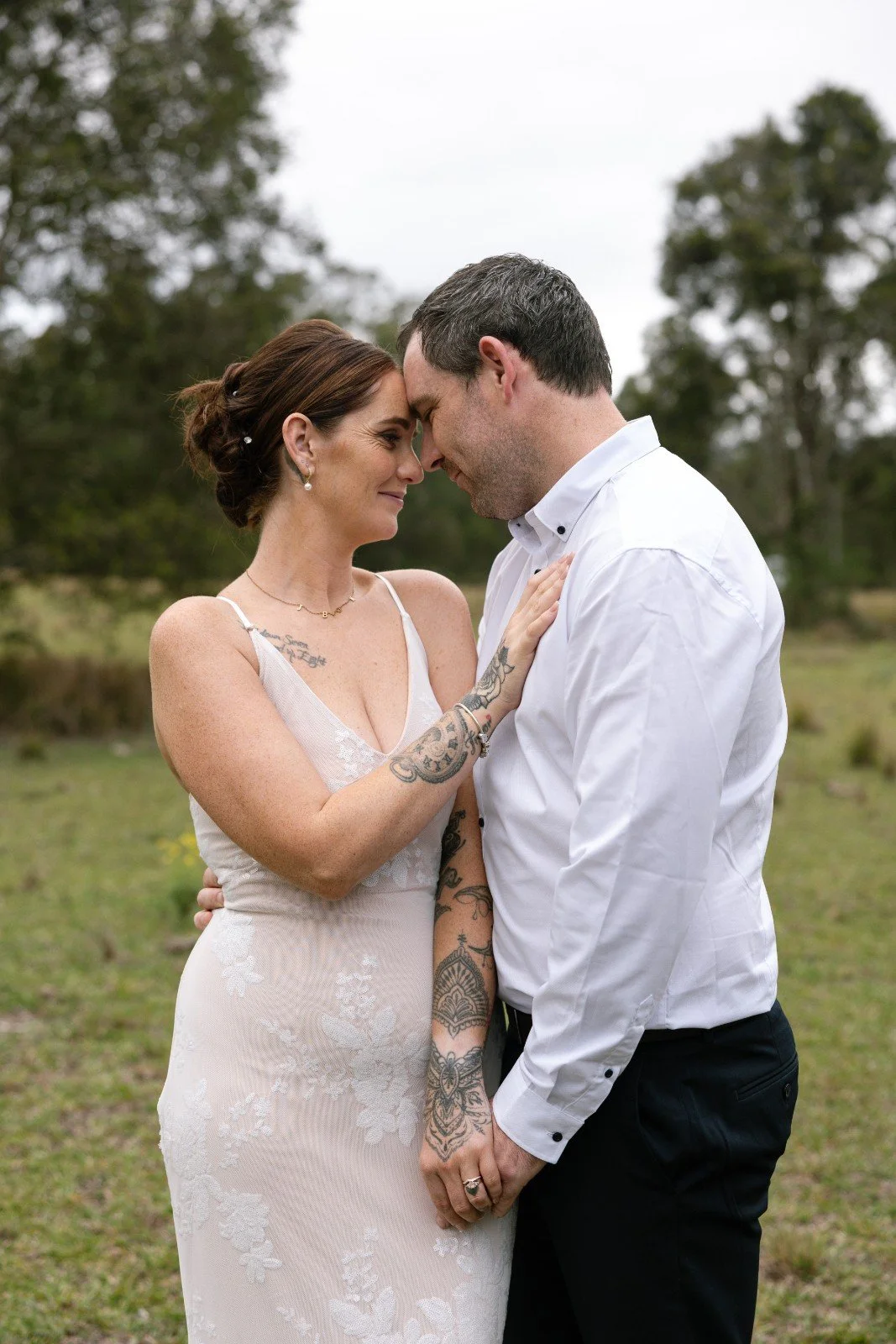 A couple touching foreheads and smiling outdoors on a cloudy day, with a woman in a white lace dress and tattoos, and a man in a white shirt and black pants.
