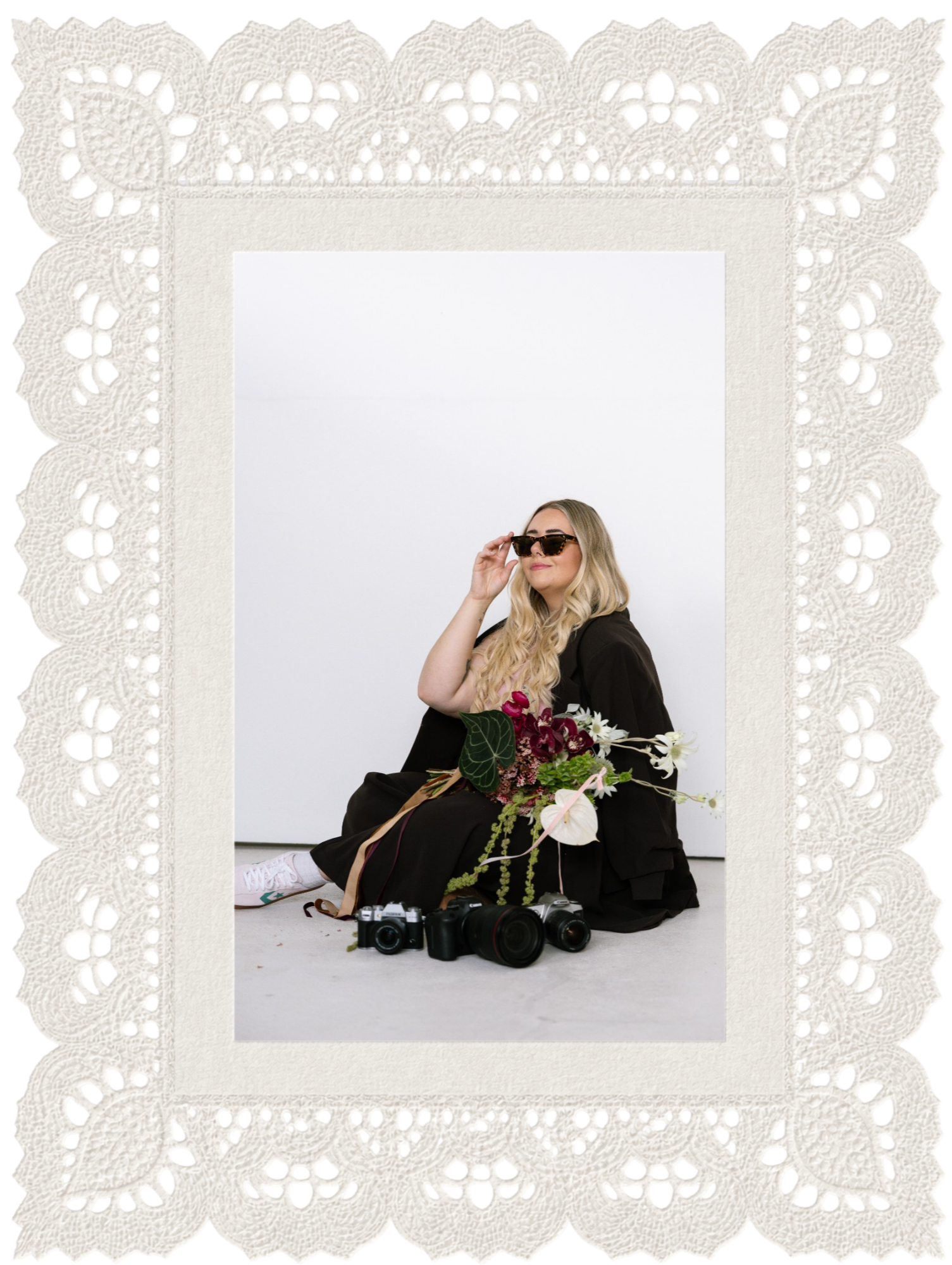 A woman sitting on the floor holding a bouquet of flowers, wearing sunglasses and a black outfit, with two cameras and a lens on the floor in front of her behind a lace-edged photo border.