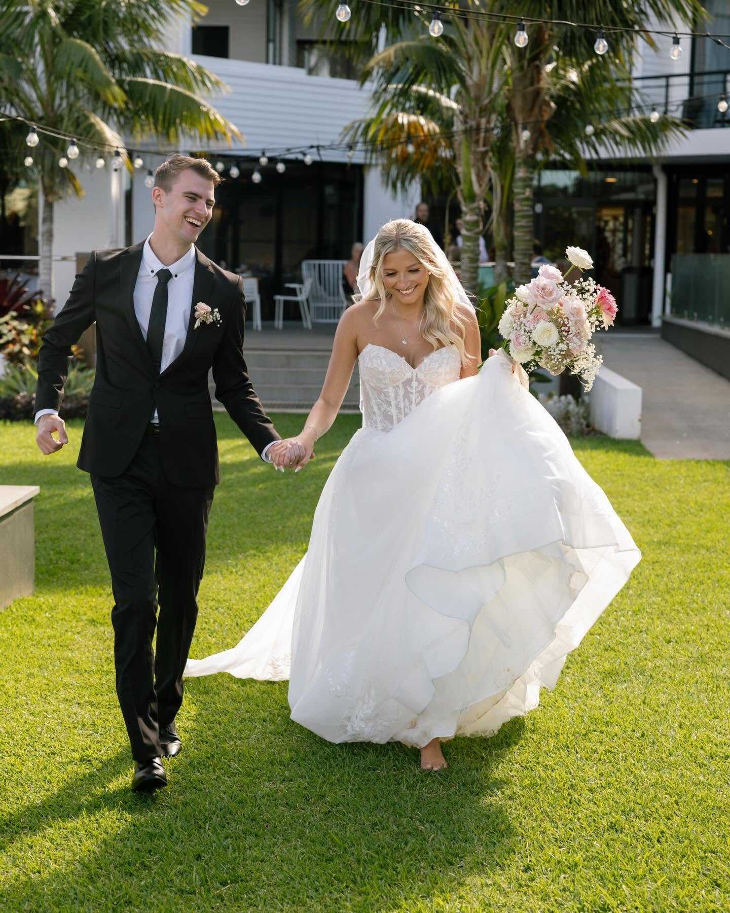 A bride and groom walking hand in hand outdoors, smiling and dressed in wedding attire, with the bride holding a bouquet of flowers.