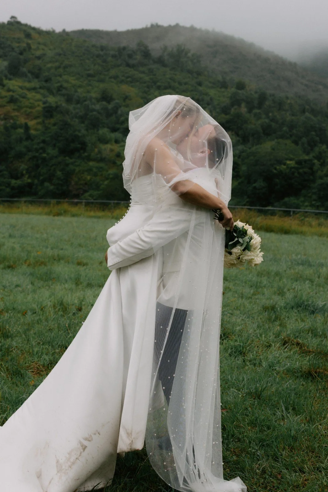A bride and groom embrace outdoors during the daytime, with the bride holding a bouquet of white flowers, and both dressed in white wedding attire, standing on a grassy field with a backdrop of foggy mountains and lush greenery.