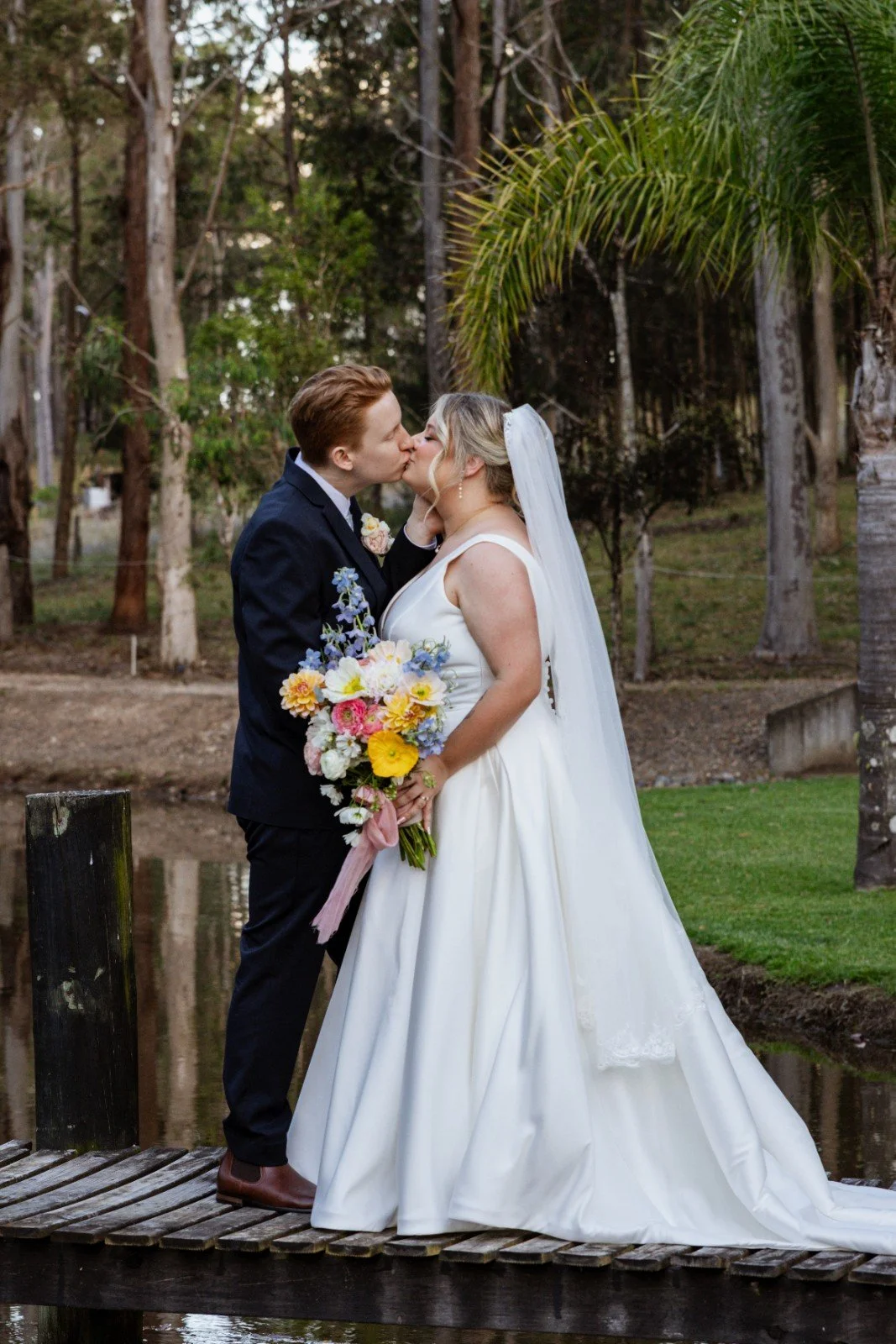 A newlywed couple shares a kiss outdoors on a wooden dock. The bride holds a colorful bouquet, and the groom wears a navy suit. Tall trees surround a pond in the background.