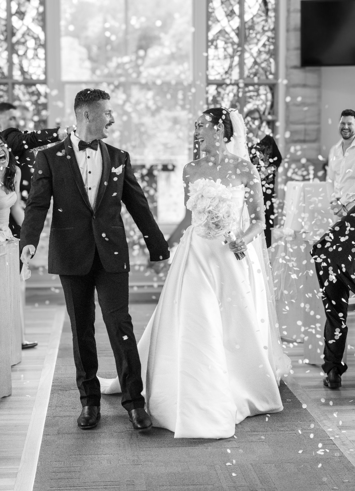A bride and groom walking hand-in-hand at their wedding, smiling joyfully, with guests throwing confetti in a wedding venue with large windows and natural light.