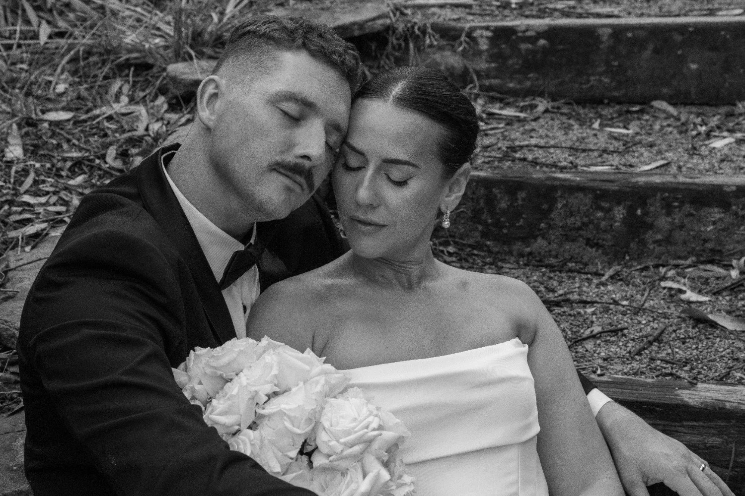 A black and white photo of a couple, the man in a tuxedo, and the woman in a strapless dress, sitting close together with their eyes closed, holding a bouquet of flowers, outdoors with wooden steps and dirt in the background.