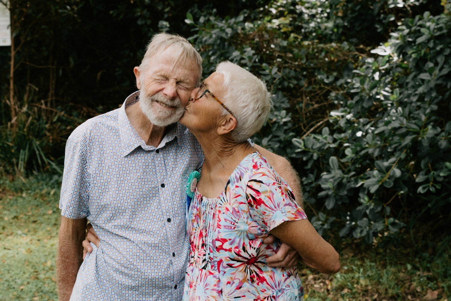 An elderly couple sharing a kiss outdoors, with greenery in the background.