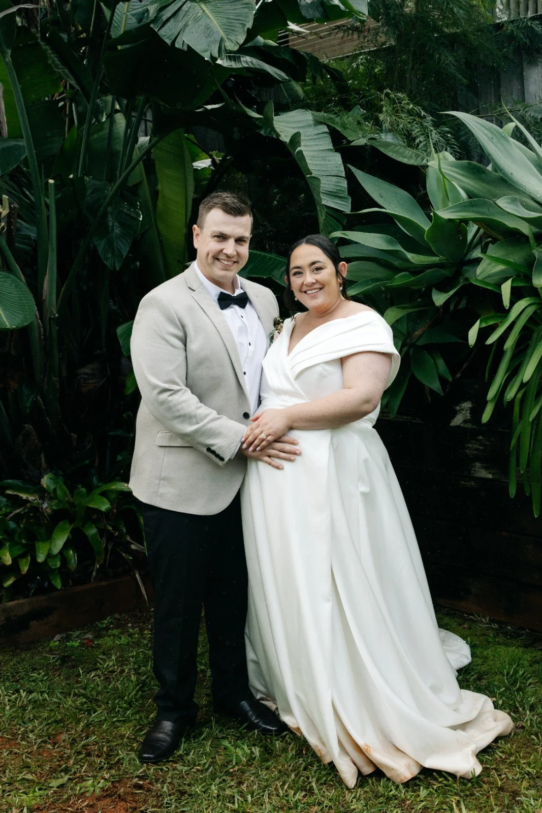 A smiling couple in wedding attire standing outdoors in front of lush green tropical plants, with the groom in a light suit and the bride in a white wedding dress.