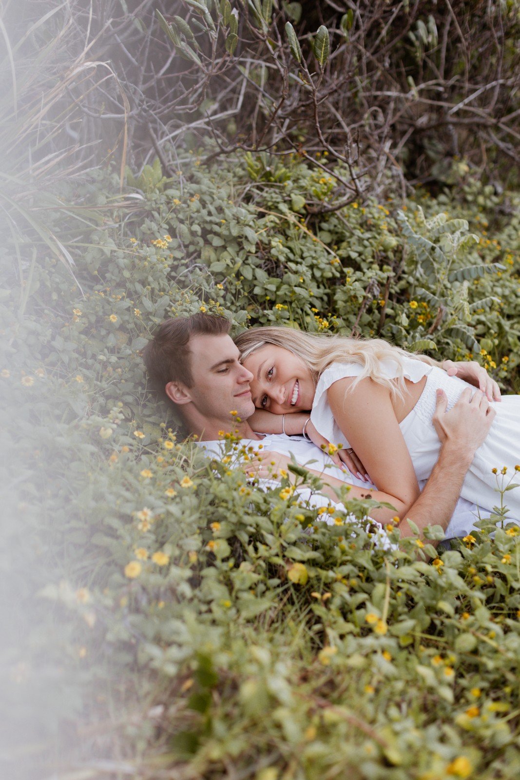 A young couple lying together in a lush green area with small yellow flowers, embracing and smiling at each other.