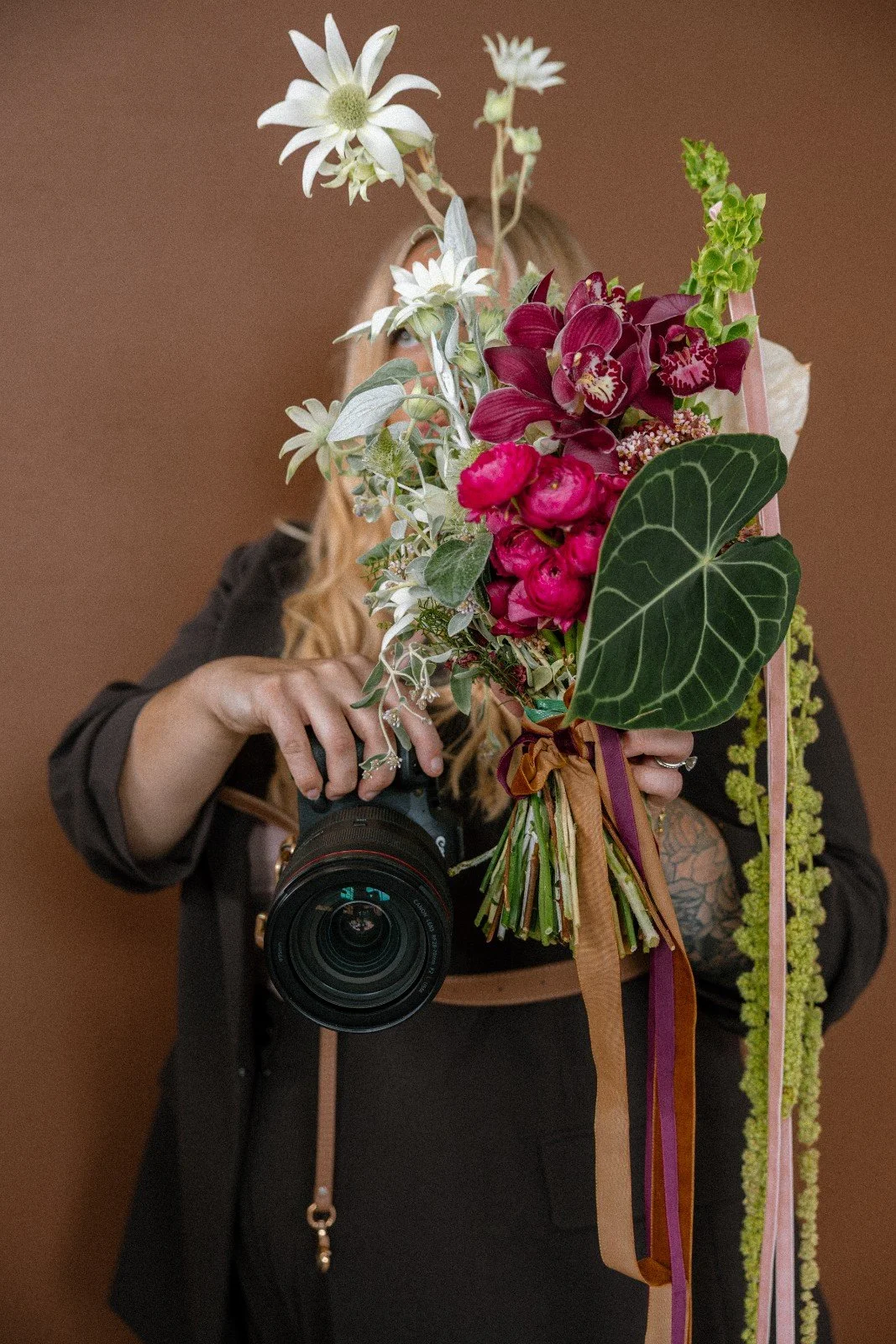 Person holding a camera and a large bouquet of various flowers, with a brown background.