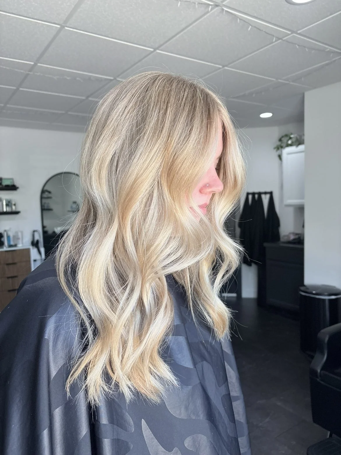 Woman with long blonde wavy hair in a salon. She is wearing a black salon cape. The background shows salon equipment and chairs.