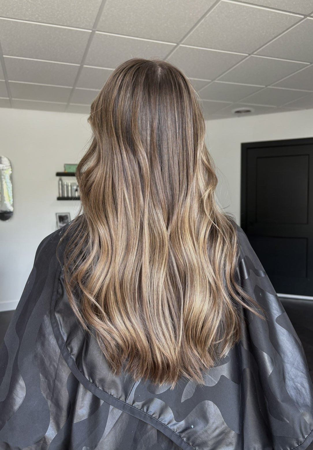 Back view of a woman with long, wavy, light brown hair with blonde highlights in a salon chair wearing a black cape.