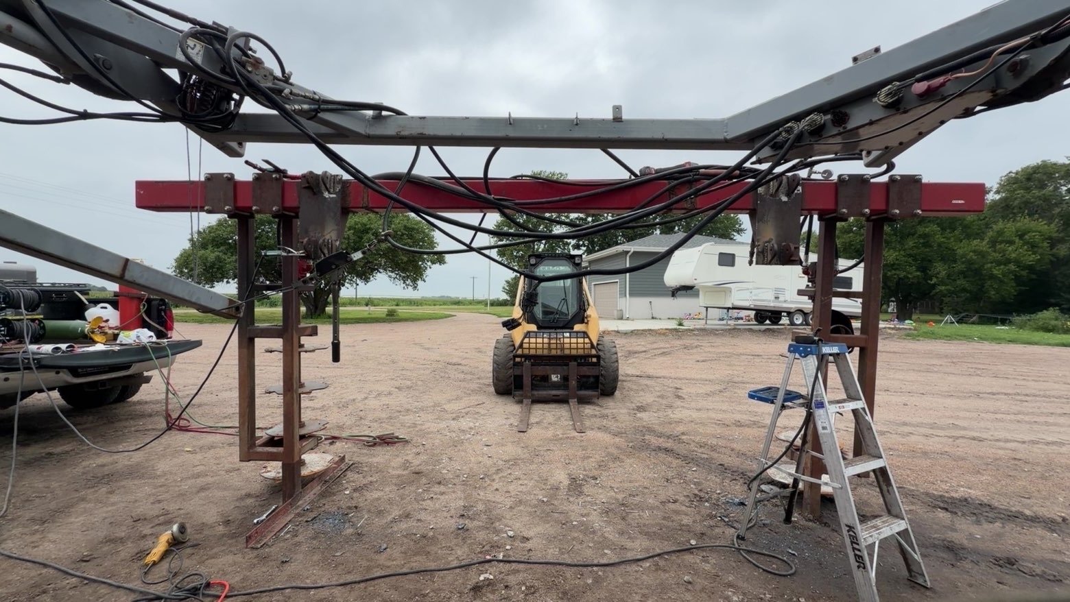 Construction site with a partially assembled metal structure, wires, a ladder, and a small construction vehicle in the background, on a dirt lot with trees and a building.