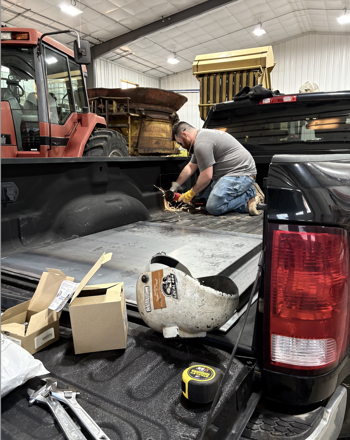 A man kneeling in the truck bed of a black pickup truck, using a grinder to cut metal, with sparks flying. There are various tools and boxes nearby inside a workshop, with a red tractor and a rusted piece of equipment in the background.