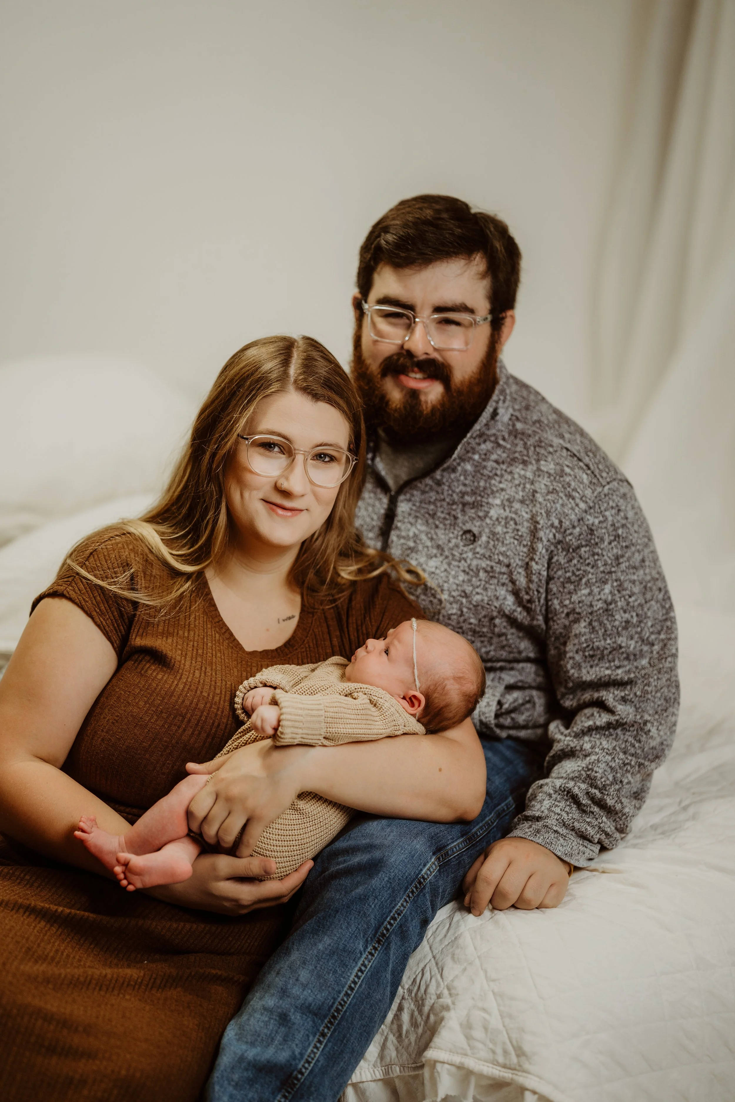 A family of four, including a woman, a man, and a newborn baby, sitting on a bed indoors. The woman is holding the baby, and the man is seated nearby. The family appears happy and comfortable.