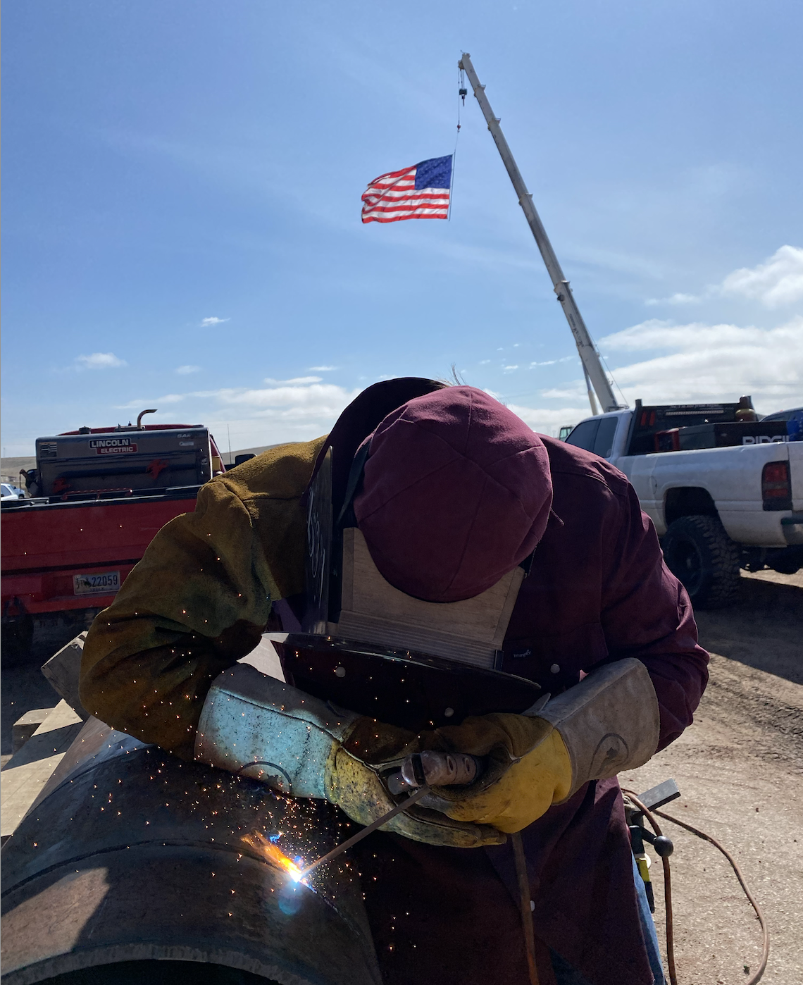 A person welding a vehicle with sparks flying, wearing protective gloves and a hood, outdoors with trucks and an American flag flying in the background.