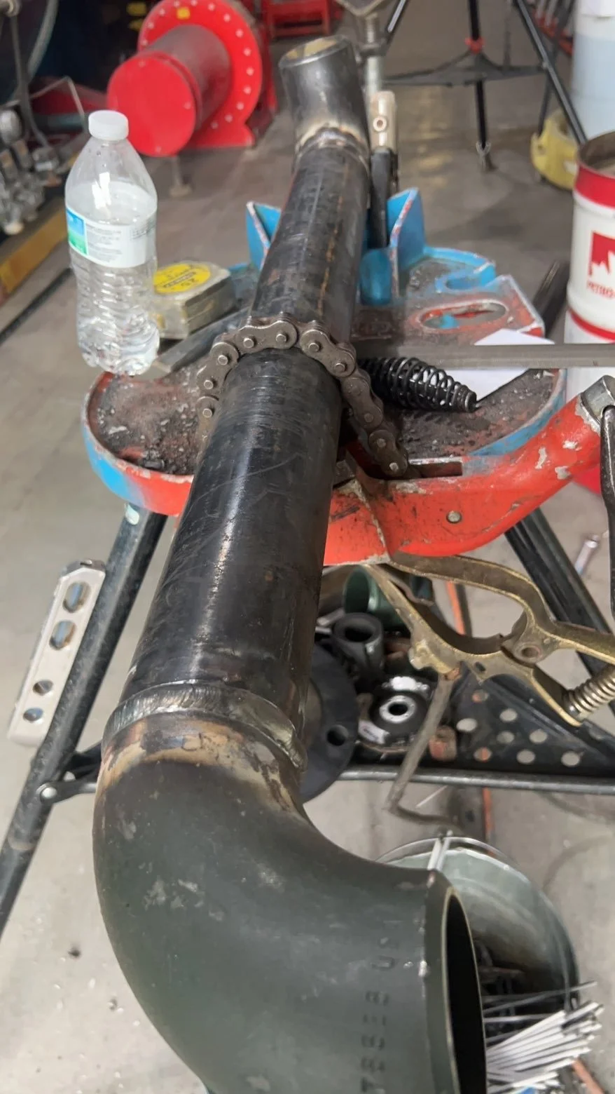 View of a large black metal pipe clamped in a workbench with various tools and items around, including a water bottle, a chain, and a spring, in a workshop setting.