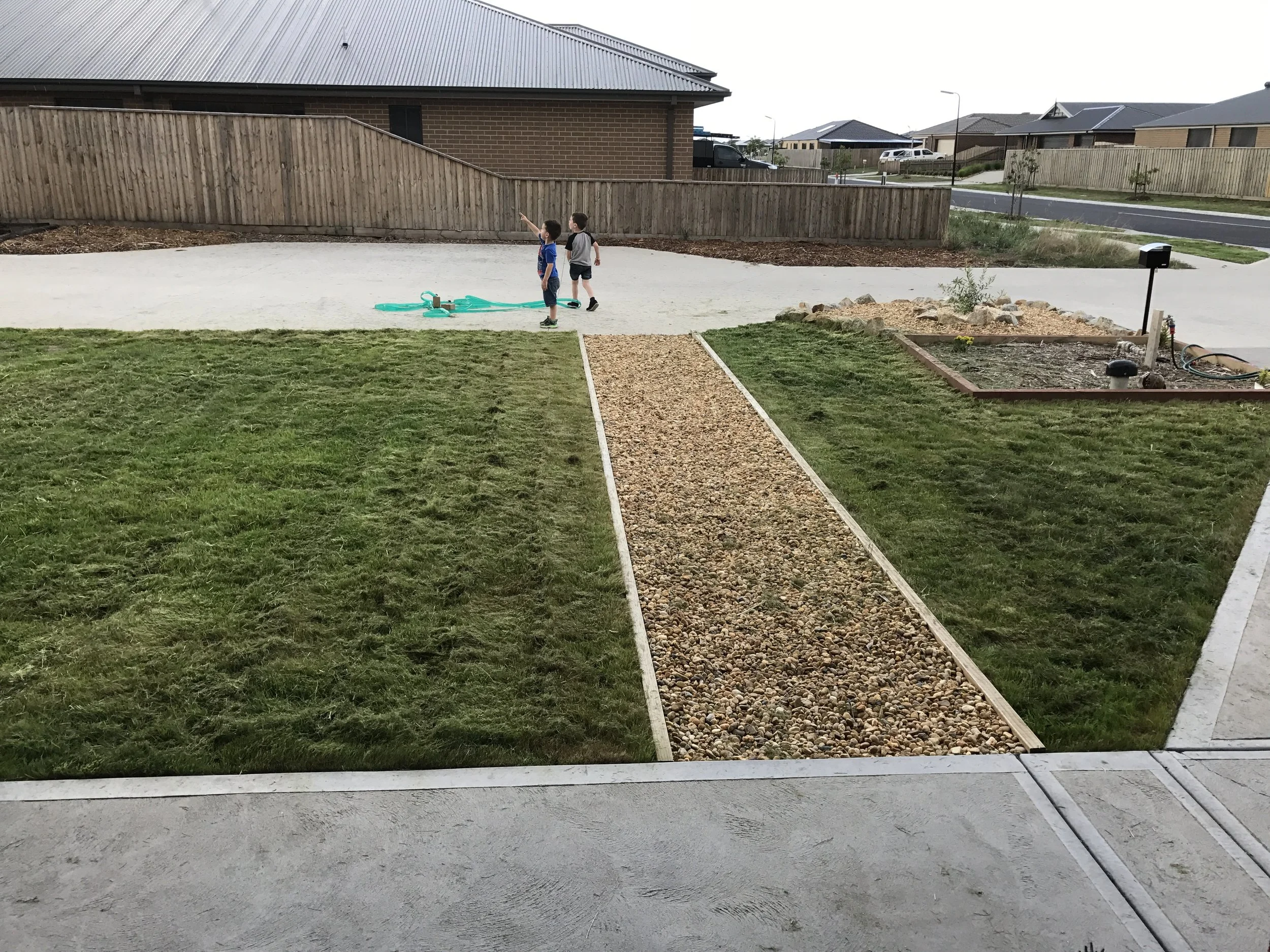 Two children playing on a concrete driveway at the back of a suburban house, with a wooden fence, a grassy lawn, and neighboring houses in the background.