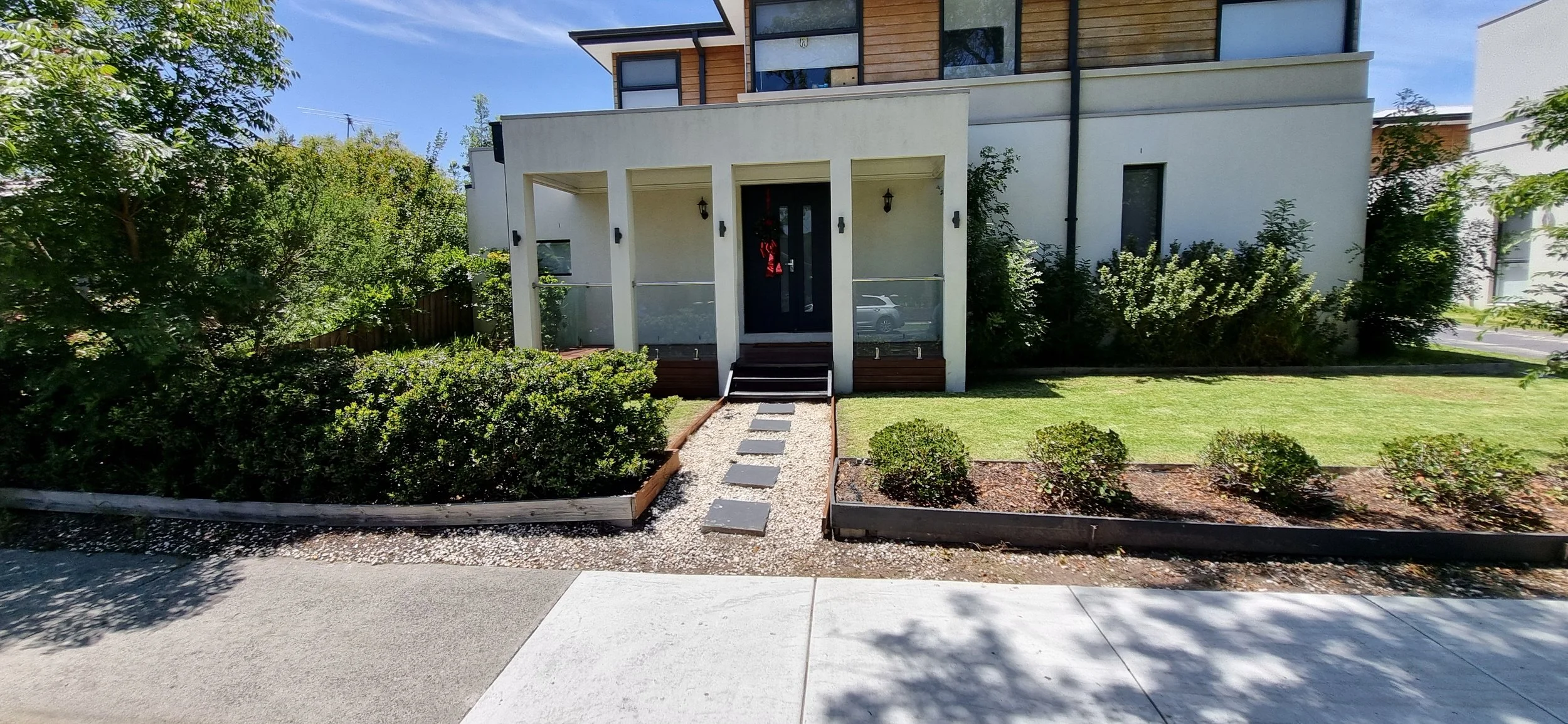 Front view of a modern house with a small front yard, stairs leading to the front door, surrounded by well-maintained bushes and trees, under a clear blue sky.