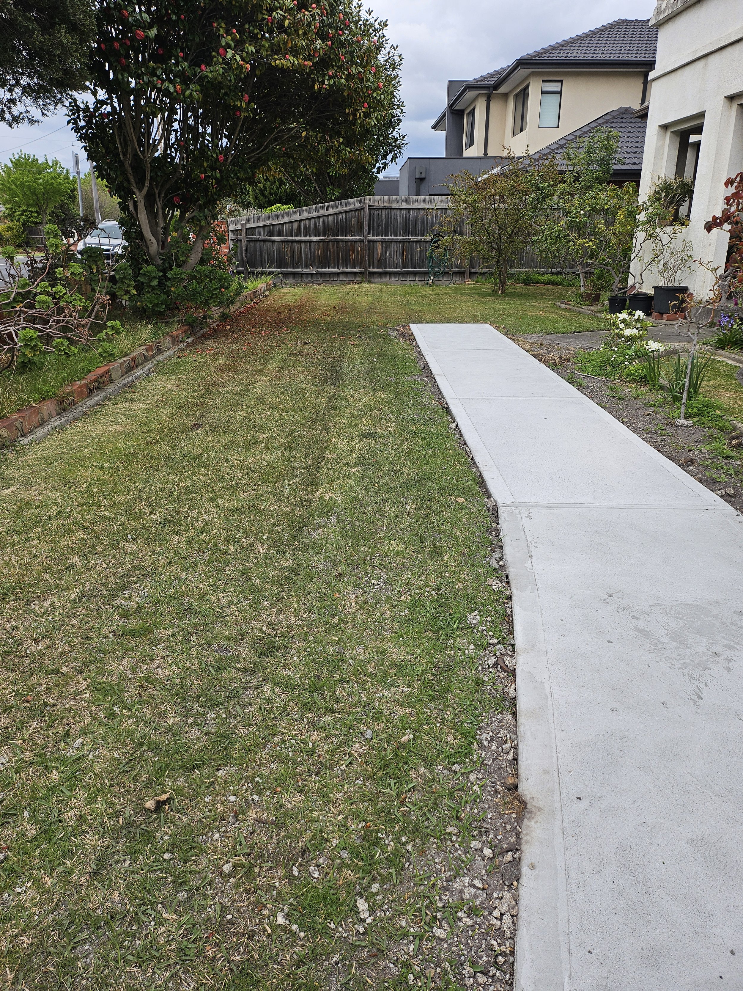 A backyard with a sidewalk, grass, trees, and a wooden fence, with neighboring houses in the background.