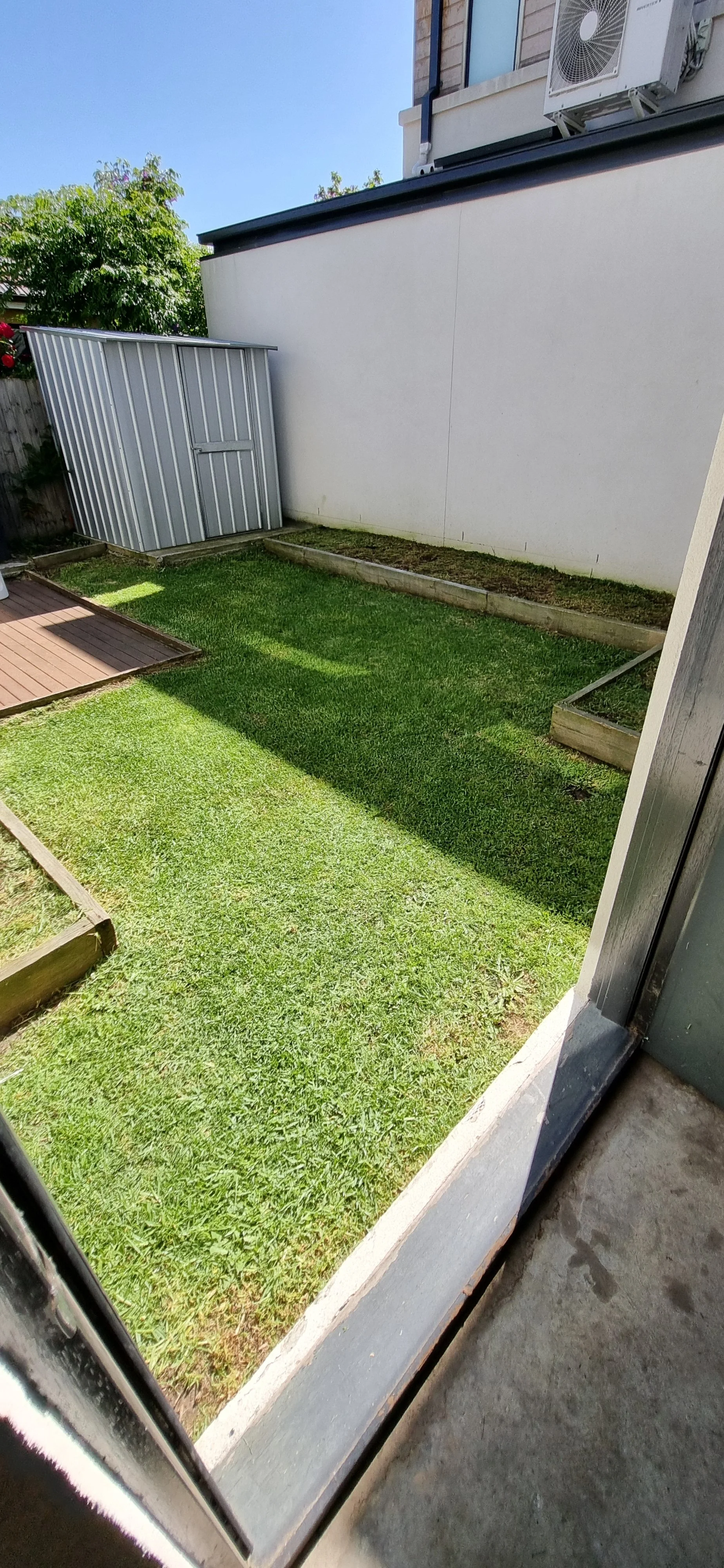 View of a small backyard with green grass, garden beds, a gray shed, and a white wall in the background, seen from inside through a sliding door.