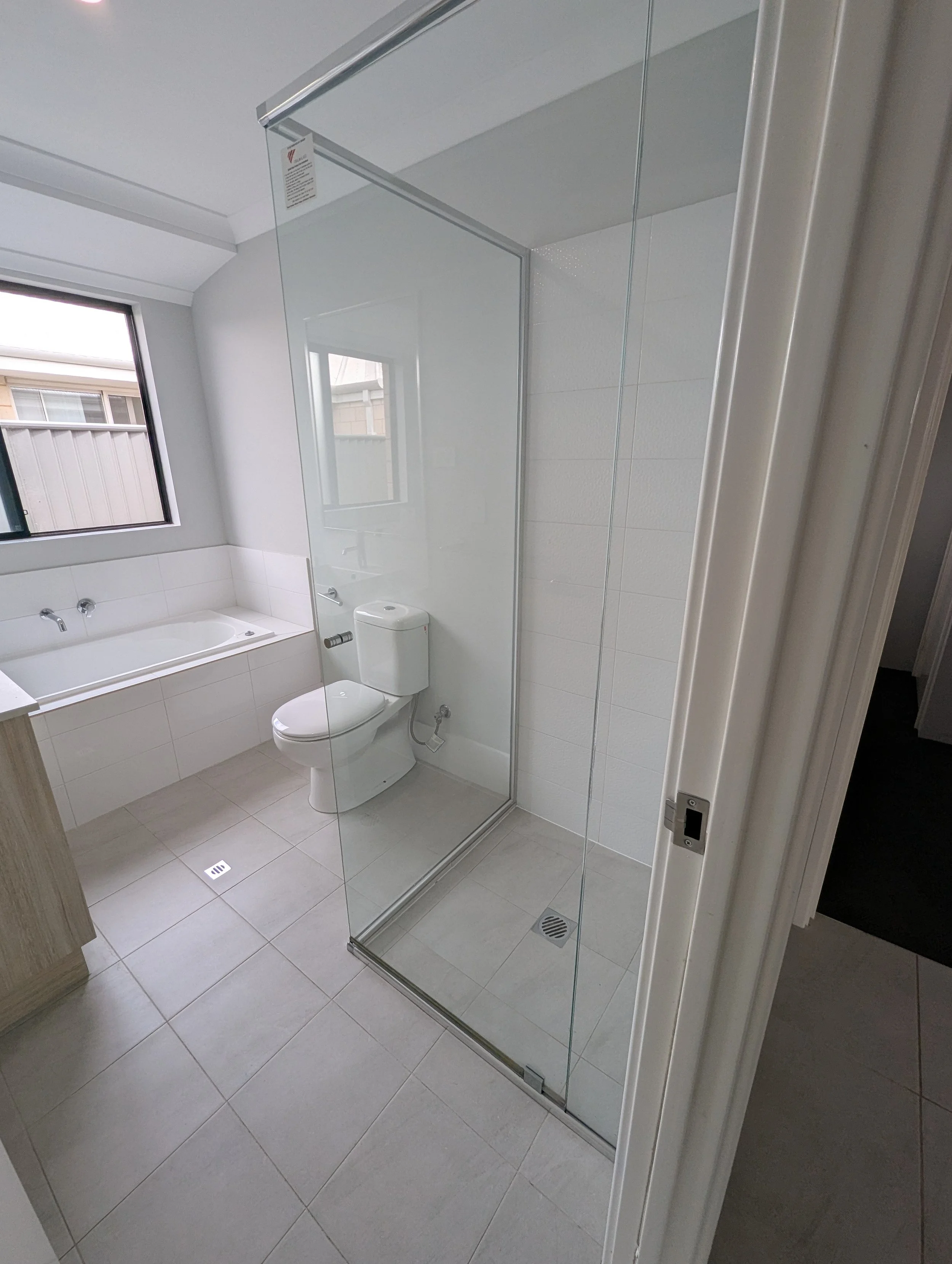 A bathroom with a bathtub, toilet, and a glass shower door. There is a window above the bathtub, and the floor and walls are tiled in white.