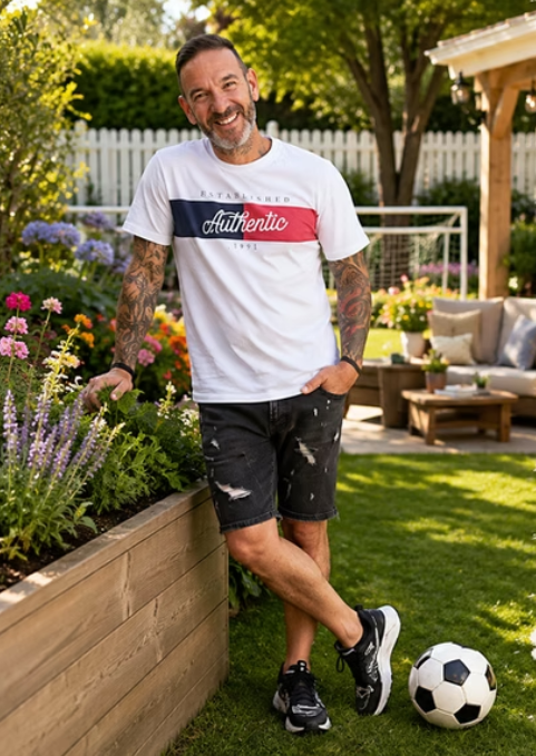 Man with gray beard and tattoos standing in a garden with colorful flowers, wearing a white T-shirt, black distressed shorts, and sneakers, smiling, with a soccer ball on the grass.